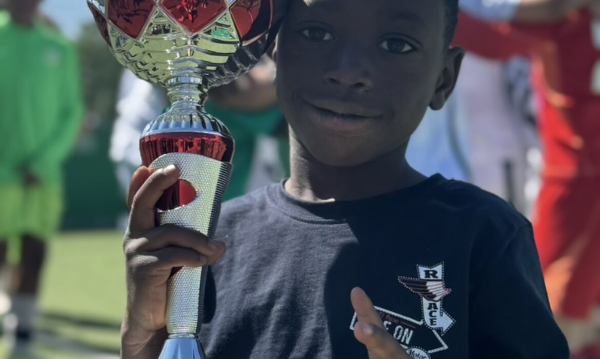Junge hält stolz einen großen silbernen Pokal mit rotem Emblem des OC-Vereins. Auf der Plakette steht: „1. Sieger U11 Intan-Cup 2025 Goldrunde“. Im Hintergrund Kinder in Fußballkleidung, sonniges Wetter und ein Sportplatz mit grünem Kunstrasen.