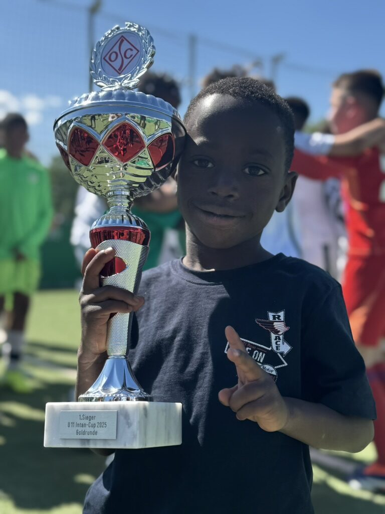 Junge hält stolz einen großen silbernen Pokal mit rotem Emblem des OC-Vereins. Auf der Plakette steht: „1. Sieger U11 Intan-Cup 2025 Goldrunde“. Im Hintergrund Kinder in Fußballkleidung, sonniges Wetter und ein Sportplatz mit grünem Kunstrasen.