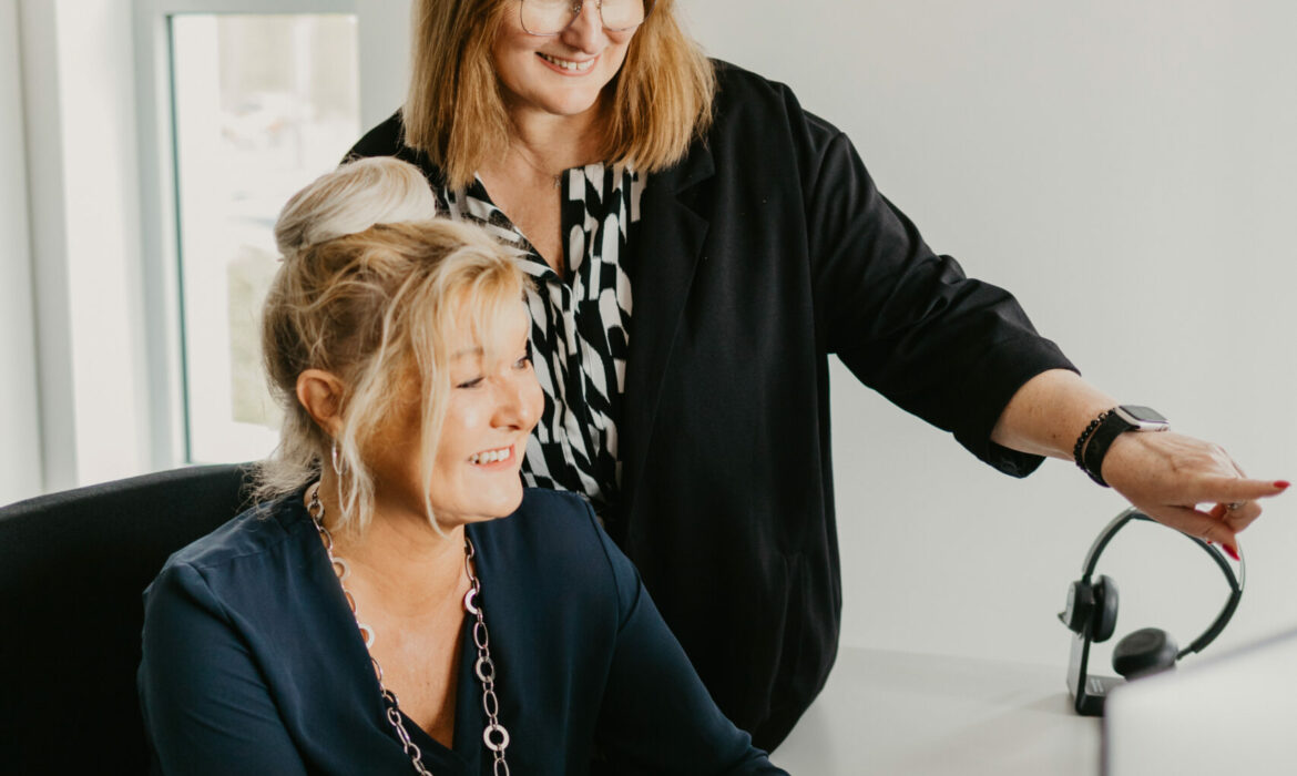 Zwei Frauen arbeiten gemeinsam an einem Computer. Eine Frau sitzt am Schreibtisch und bedient konzentriert die Maus. Die andere Frau steht lächelnd daneben und schaut auf den Bildschirm. Beide wirken konzentriert und im Austausch, die Szene vermittelt Teamarbeit in einer modernen Büroumgebung. Im Vordergrund liegen bunte Zeitschriften, die den Print-Bezug unterstreichen.