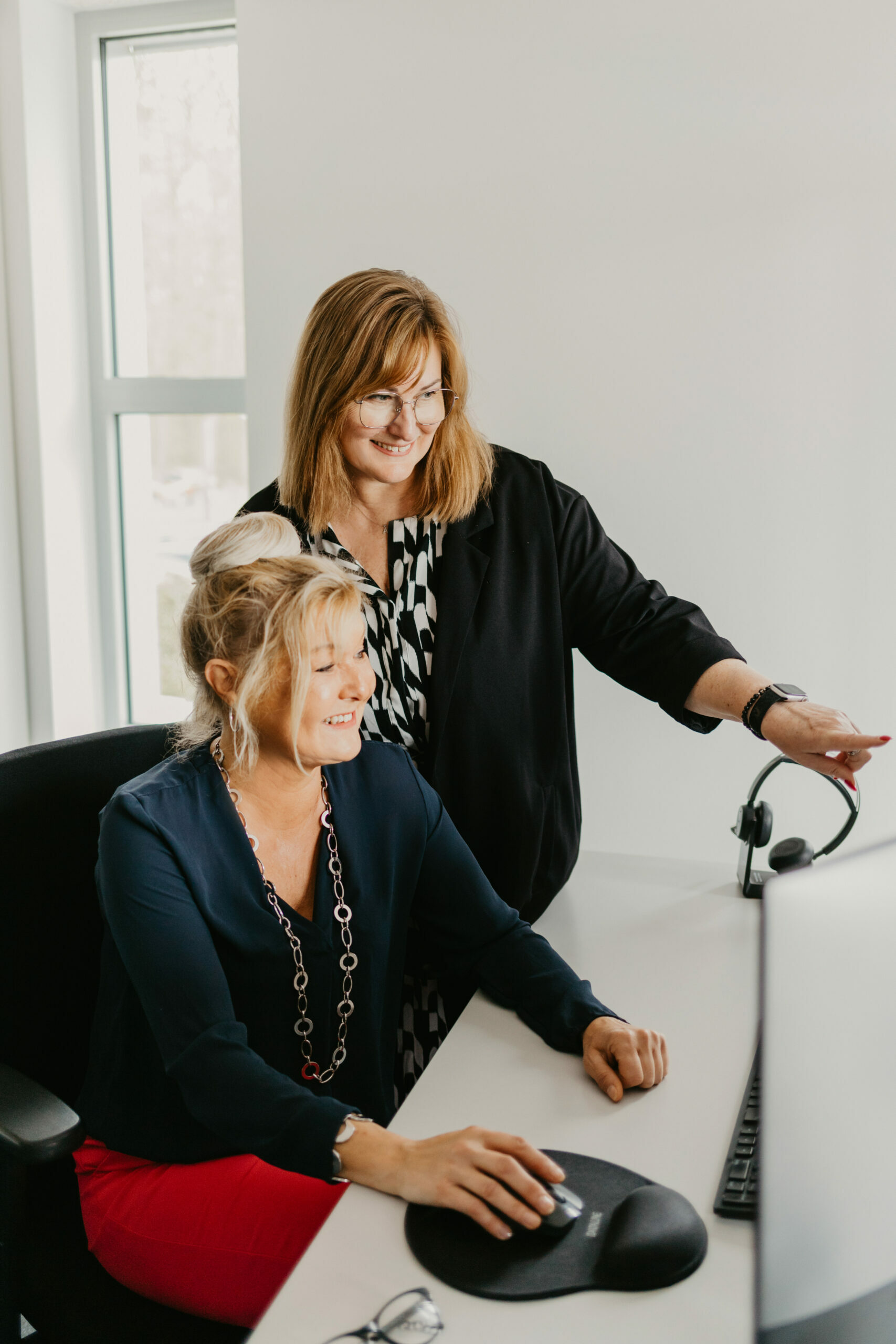 Zwei Frauen arbeiten gemeinsam an einem Computer. Eine Frau sitzt am Schreibtisch und bedient konzentriert die Maus. Die andere Frau steht lächelnd daneben und schaut auf den Bildschirm. Beide wirken konzentriert und im Austausch, die Szene vermittelt Teamarbeit in einer modernen Büroumgebung. Im Vordergrund liegen bunte Zeitschriften, die den Print-Bezug unterstreichen.