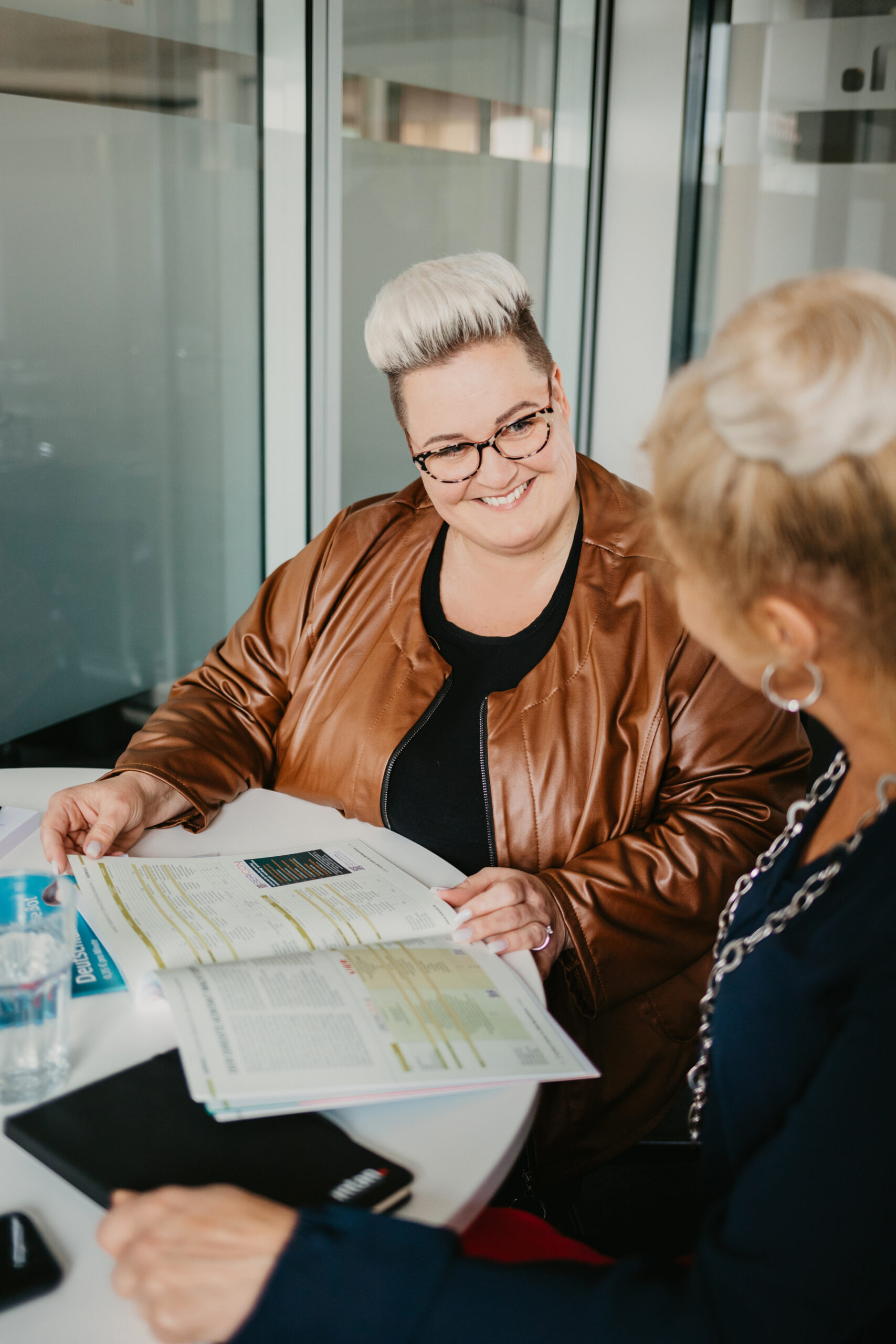 Dieses Bild zeigt ein professionelles, freundliches Gespräch in einer modernen Büroumgebung. Die Person mit heller Kurzhaarfrisur und brauner Jacke lächelt herzlich und zeigt ihrer Gesprächspartnerin etwas in einer aufgeschlagenen Broschüre oder einem Magazin. Beide wirken konzentriert und zugewandt. Die Atmosphäre vermittelt Vertrauen, Offenheit und Zusammenarbeit – ideal für interne Kommunikation, Beratung oder Teamwork-Visualisierungen.