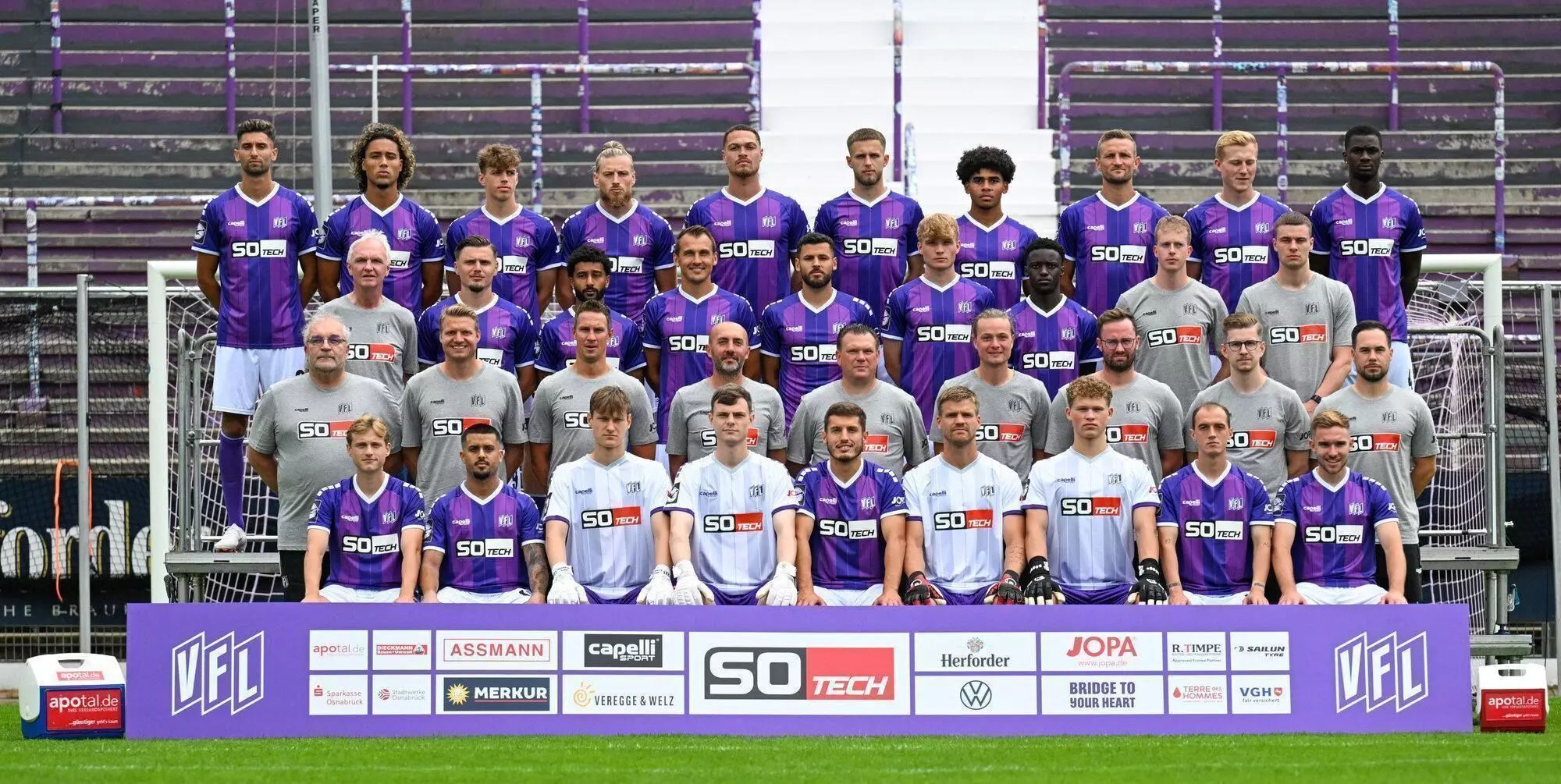 Mannschaftsfoto eines Fußballteams im Stadion. Etwa 35 Personen stehen und sitzen in mehreren Reihen vor leeren Tribünen. Die Spieler tragen überwiegend lila Trikots mit weißen Hosen, einige weiße Trikots. Davor steht eine Werbebande mit Logos und dem VfL-Emblem.