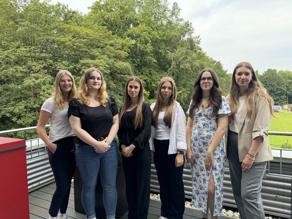 Sechs junge Frauen stehen nebeneinander auf einer Terrasse vor einer grünen Waldkulisse. Sie schauen lächelnd in die Kamera und tragen sommerliche Kleidung in hellen und dunklen Farbtönen.