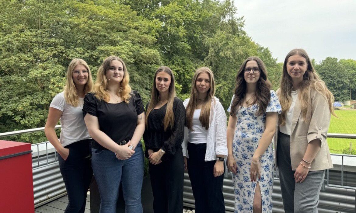Sechs junge Frauen stehen nebeneinander auf einer Terrasse vor einer grünen Waldkulisse. Sie schauen lächelnd in die Kamera und tragen sommerliche Kleidung in hellen und dunklen Farbtönen.