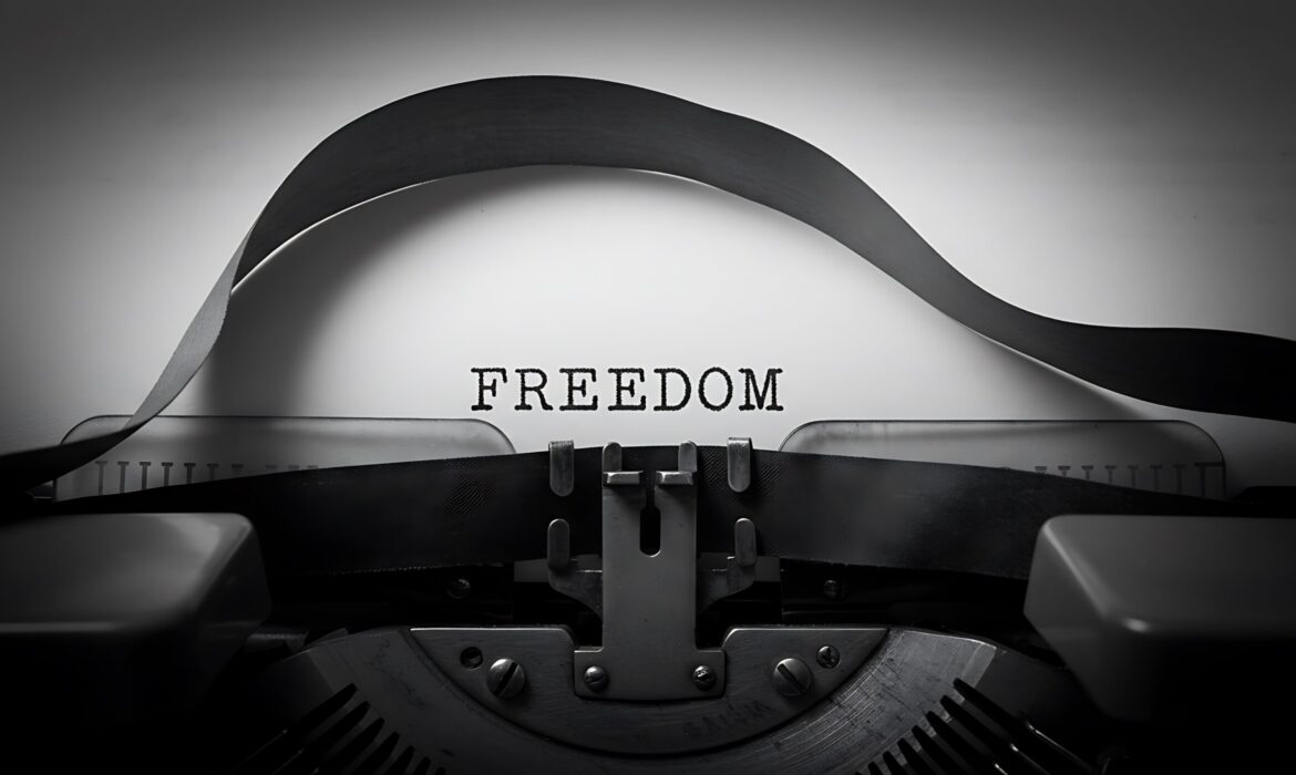 Eine Nahaufnahme einer Schreibmaschine, auf deren Papier das Wort „FREEDOM“ in Großbuchstaben getippt ist. Das Bild ist in Schwarz-Weiß gehalten und wirkt symbolisch und kontrastreich.