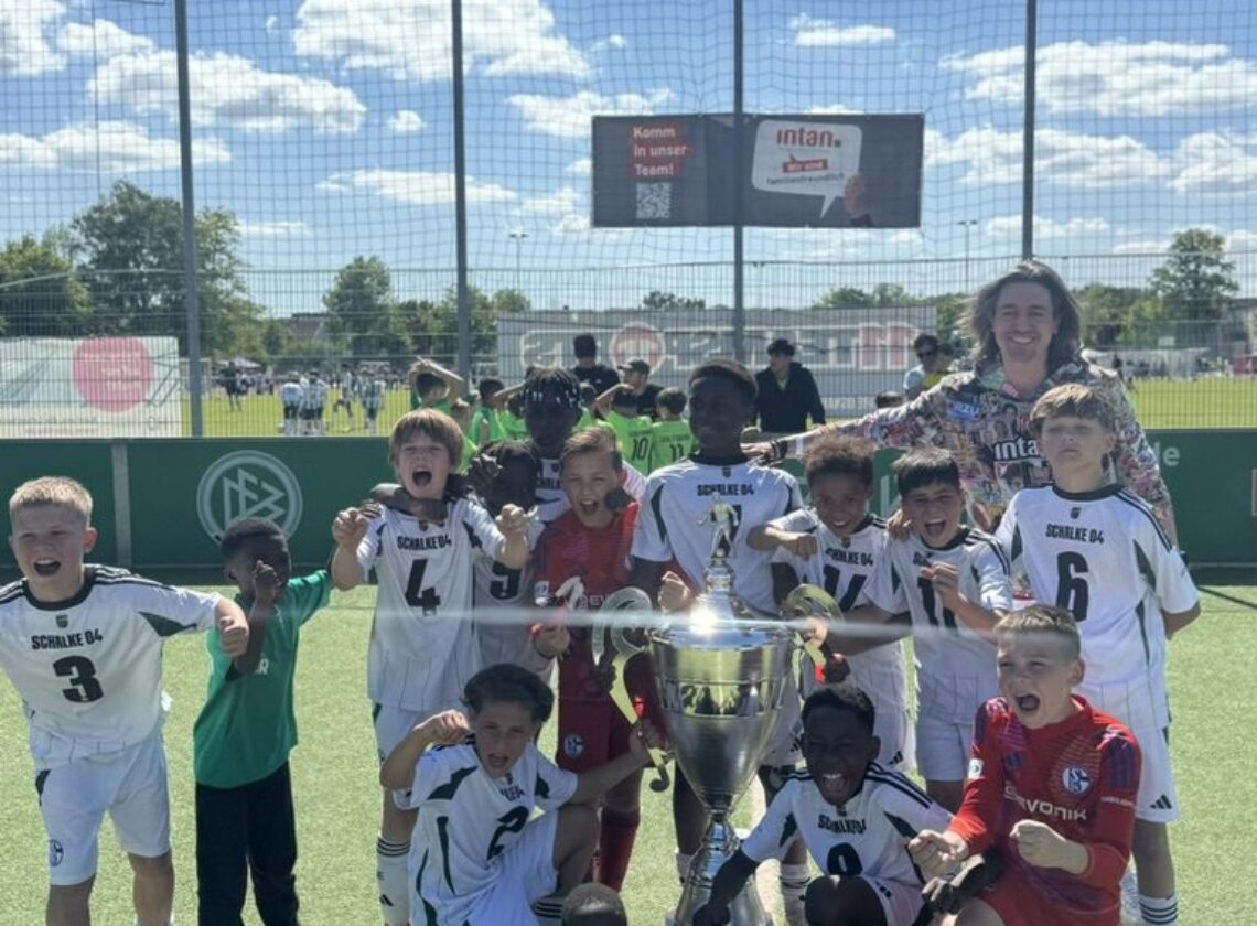 Kinderfußballmannschaft jubelt gemeinsam mit einer Trainerin auf einem Sportplatz bei Sonnenschein. Die Kinder tragen überwiegend weiße Trikots mit dem Aufdruck „Schalke 04“ und versammeln sich um einen großen silbernen Pokal. Im Hintergrund ist ein Spielfeld mit weiteren Spielern und ein Werbebanner der intan group zu sehen.