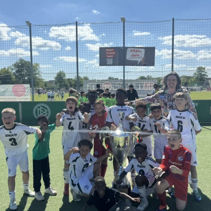 Kinderfußballmannschaft jubelt gemeinsam mit einer Trainerin auf einem Sportplatz bei Sonnenschein. Die Kinder tragen überwiegend weiße Trikots mit dem Aufdruck „Schalke 04“ und versammeln sich um einen großen silbernen Pokal. Im Hintergrund ist ein Spielfeld mit weiteren Spielern und ein Werbebanner der intan group zu sehen.