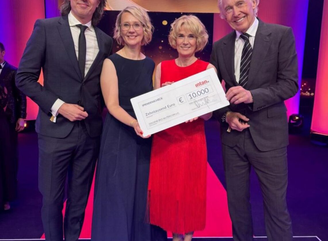 Vier festlich gekleidete Personen stehen auf einer Bühne bei der Preisverleihung „Die Goldene Bild der Frau 2024“. Sie halten gemeinsam einen großen symbolischen Spendenscheck der intan group über 10.000 Euro. Im Hintergrund leuchten pinke und goldene Lichteffekte, auf der Bühne ist das Logo der Veranstaltung zu sehen.