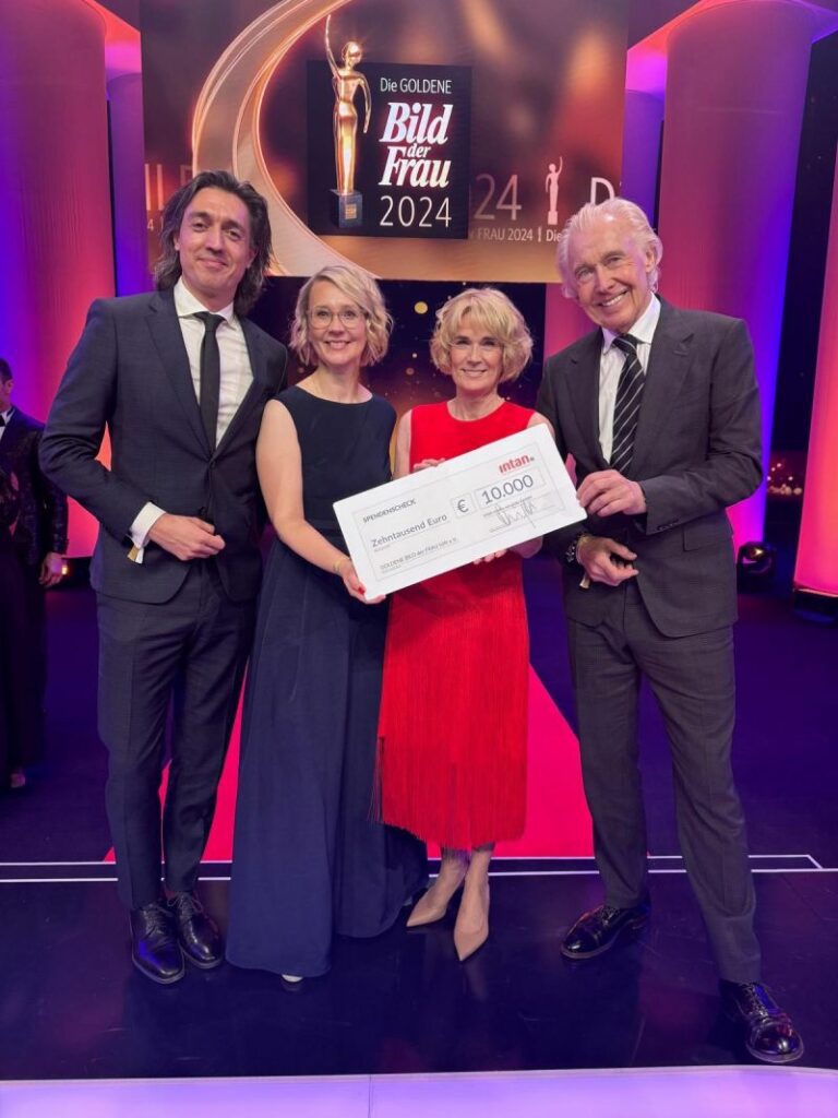 Vier festlich gekleidete Personen stehen auf einer Bühne bei der Preisverleihung „Die Goldene Bild der Frau 2024“. Sie halten gemeinsam einen großen symbolischen Spendenscheck der intan group über 10.000 Euro. Im Hintergrund leuchten pinke und goldene Lichteffekte, auf der Bühne ist das Logo der Veranstaltung zu sehen.