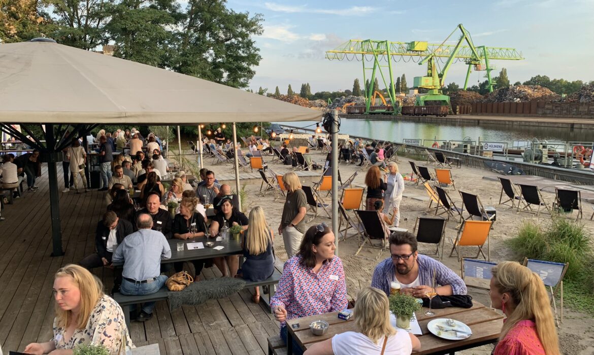 Menschen sitzen an Holztischen unter einem großen Sonnenschirm und auf Liegestühlen am Sandstrand eines Hafenareals. Im Hintergrund ein Fluss mit grün lackierten Hafenkränen und Schrottbergen. Die Stimmung ist sommerlich und gesellig.