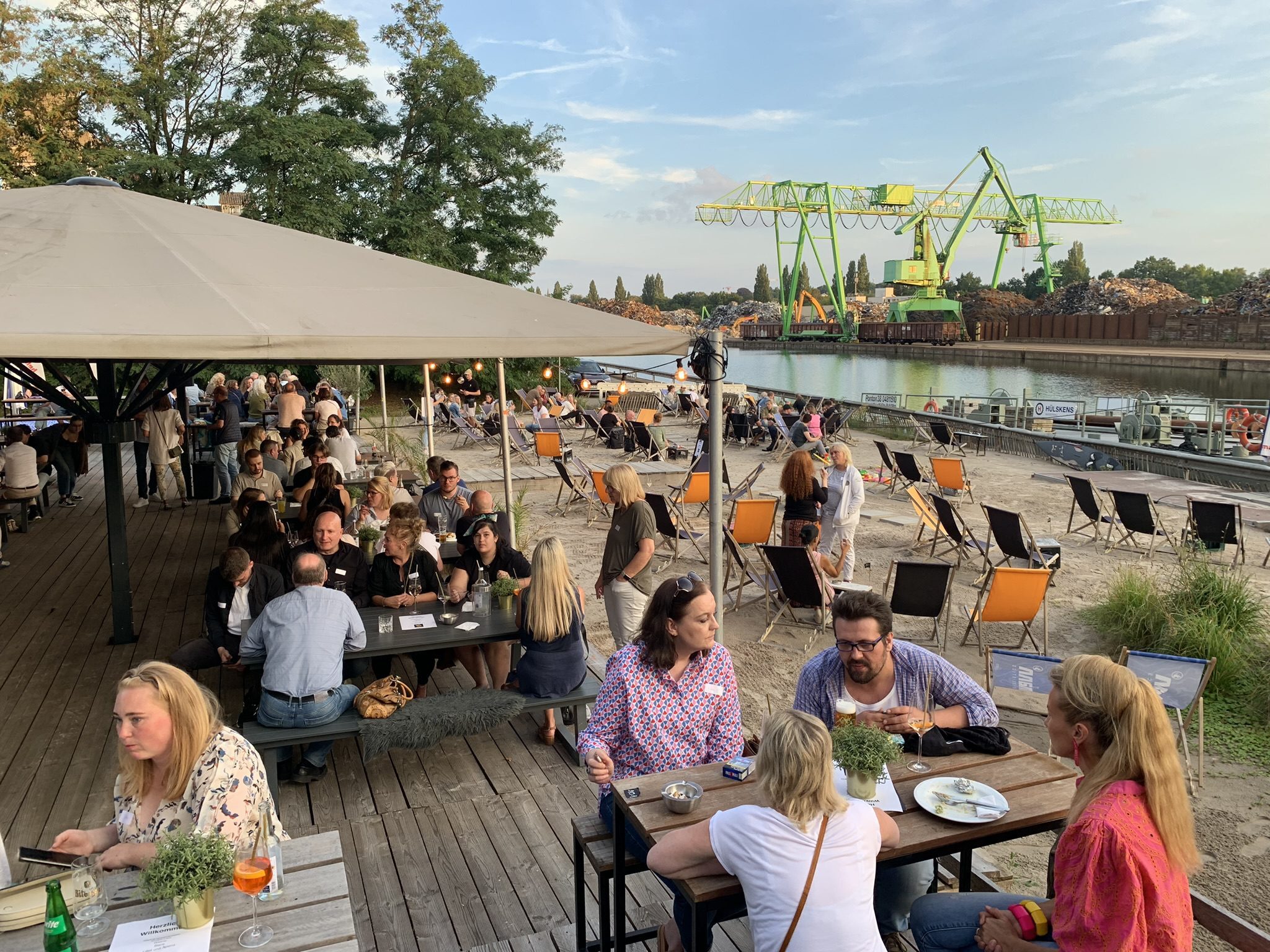 Menschen sitzen an Holztischen unter einem großen Sonnenschirm und auf Liegestühlen am Sandstrand eines Hafenareals. Im Hintergrund ein Fluss mit grün lackierten Hafenkränen und Schrottbergen. Die Stimmung ist sommerlich und gesellig.