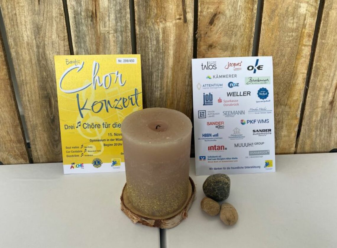 Zwei Plakate stehen nebeneinander vor einer Holzrückwand, dazwischen eine beige Kerze mit goldenen Glitzereffekten auf einem Holzuntersetzer. Links ein gelbes Plakat zum Benefiz-Chorkonzert am 15. November, rechts ein Sponsorenplakat mit zahlreichen Firmenlogos. Davor liegen vier kleine Steine und Nüsse.