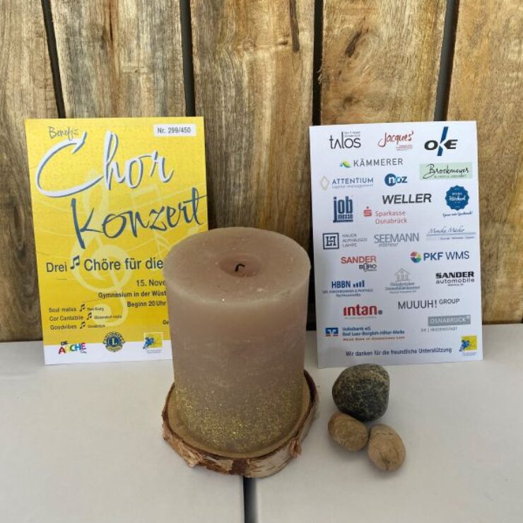 Zwei Plakate stehen nebeneinander vor einer Holzrückwand, dazwischen eine beige Kerze mit goldenen Glitzereffekten auf einem Holzuntersetzer. Links ein gelbes Plakat zum Benefiz-Chorkonzert am 15. November, rechts ein Sponsorenplakat mit zahlreichen Firmenlogos. Davor liegen vier kleine Steine und Nüsse.