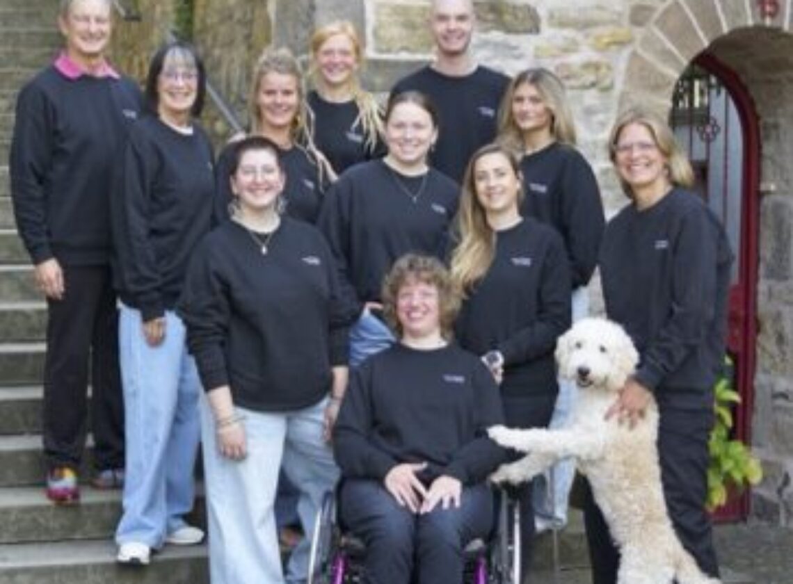 Gruppenfoto von elf Personen in schwarzer Kleidung, aufgenommen vor einem historischen Sandsteingebäude mit Treppe. Eine Person sitzt im Rollstuhl, ein weißer Hund steht mit den Vorderpfoten auf den Beinen einer Person. Die Stimmung ist freundlich und offen.