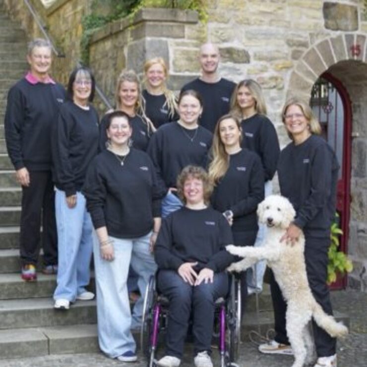 Gruppenfoto von elf Personen in schwarzer Kleidung, aufgenommen vor einem historischen Sandsteingebäude mit Treppe. Eine Person sitzt im Rollstuhl, ein weißer Hund steht mit den Vorderpfoten auf den Beinen einer Person. Die Stimmung ist freundlich und offen.