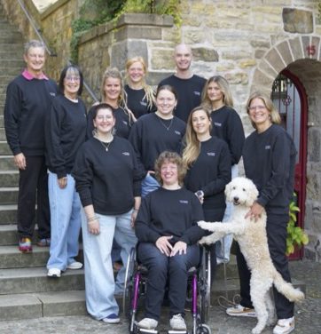 Gruppenfoto von elf Personen in schwarzer Kleidung, aufgenommen vor einem historischen Sandsteingebäude mit Treppe. Eine Person sitzt im Rollstuhl, ein weißer Hund steht mit den Vorderpfoten auf den Beinen einer Person. Die Stimmung ist freundlich und offen.