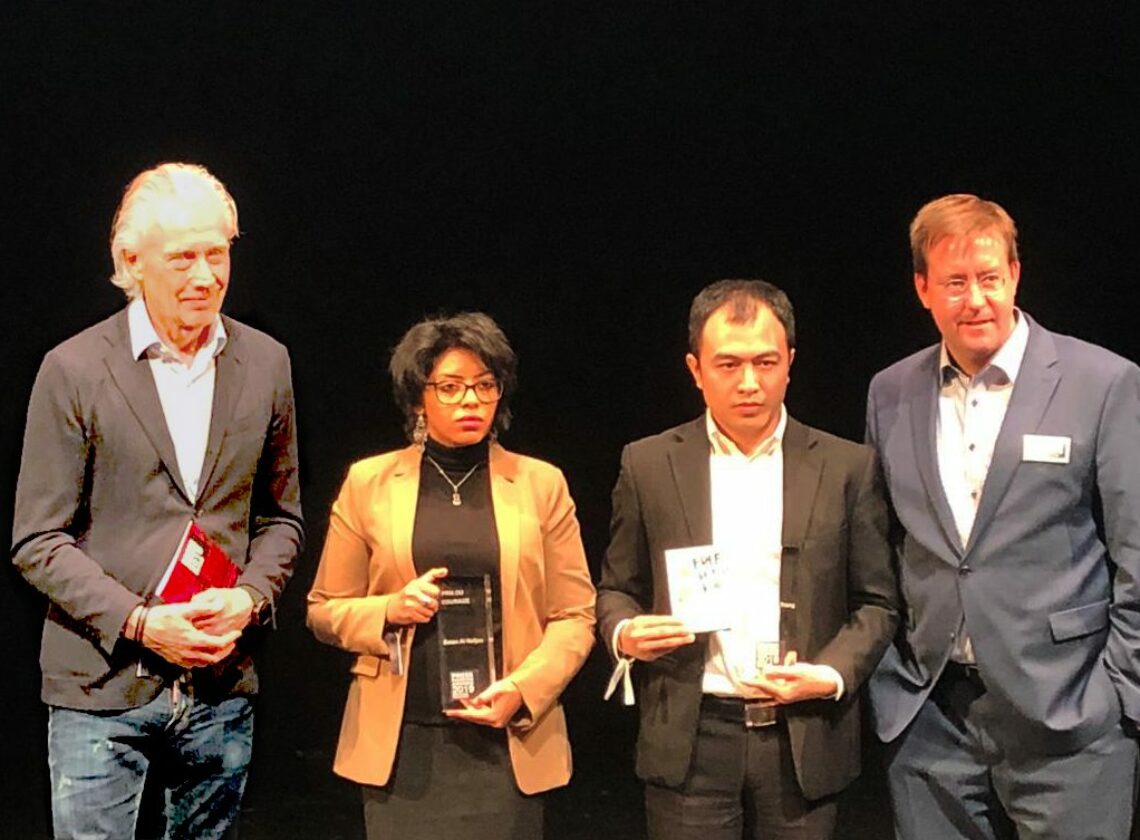 Vier Personen stehen nebeneinander auf einer Bühne vor schwarzem Hintergrund. Zwei von ihnen halten Glastrophäen, eine weitere Person hält ein Buch und ein Zertifikat. Alle sind formell gekleidet, die Szene wirkt wie eine Preisverleihung oder Ehrung.