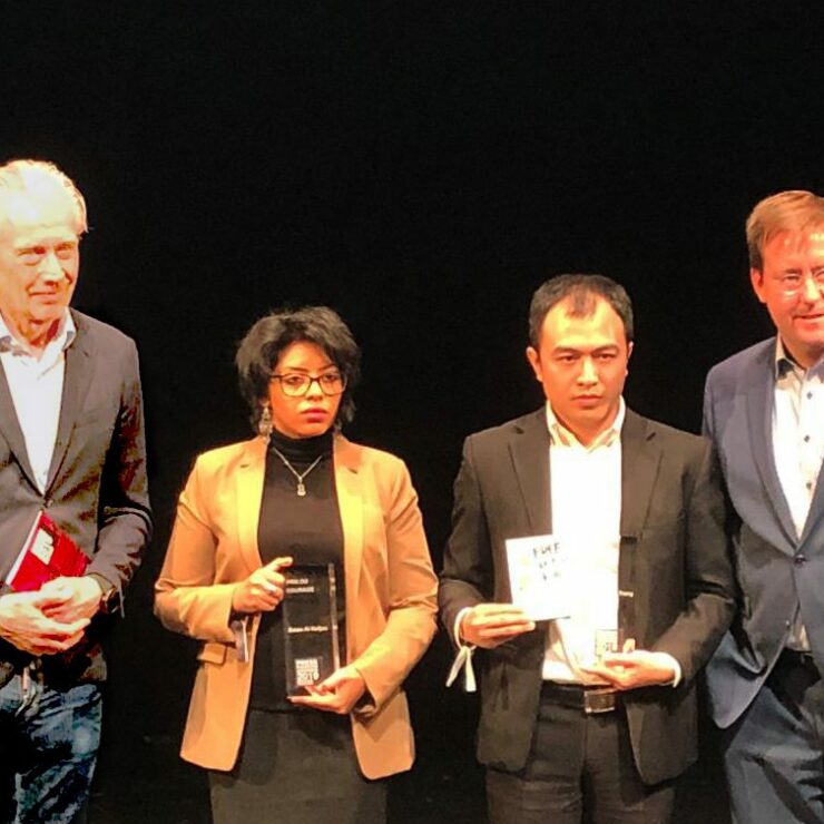 Vier Personen stehen nebeneinander auf einer Bühne vor schwarzem Hintergrund. Zwei von ihnen halten Glastrophäen, eine weitere Person hält ein Buch und ein Zertifikat. Alle sind formell gekleidet, die Szene wirkt wie eine Preisverleihung oder Ehrung.