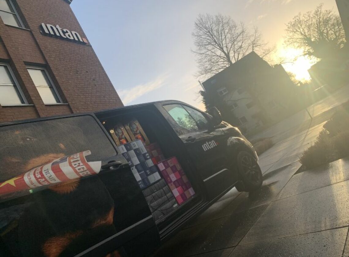 Ein schwarzer Lieferwagen mit intan-Logo steht vor einem Bürogebäude mit ebenfalls sichtbarem intan-Schriftzug. Die Schiebetür des Wagens ist geöffnet, im Inneren sind bunte Geschenkboxen und Zeitschriftenrollen zu sehen. Die Szene spielt bei Sonnenaufgang oder -untergang, mit tiefstehender Sonne im Hintergrund.