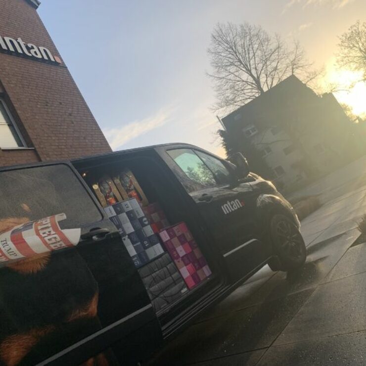 Ein schwarzer Lieferwagen mit intan-Logo steht vor einem Bürogebäude mit ebenfalls sichtbarem intan-Schriftzug. Die Schiebetür des Wagens ist geöffnet, im Inneren sind bunte Geschenkboxen und Zeitschriftenrollen zu sehen. Die Szene spielt bei Sonnenaufgang oder -untergang, mit tiefstehender Sonne im Hintergrund.