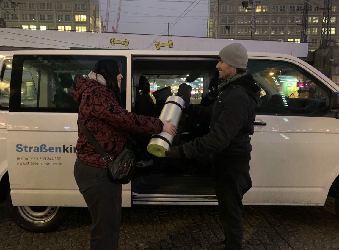 Zwei Personen stehen bei Dunkelheit vor einem weißen Kleinbus mit der Aufschrift „Straßenkinder e. V.“. Eine Person überreicht der anderen eine zusammengerollte Isomatte mit Schlafsack. Im Hintergrund sind beleuchtete Gebäude und Straßenlichter einer Stadt zu sehen.