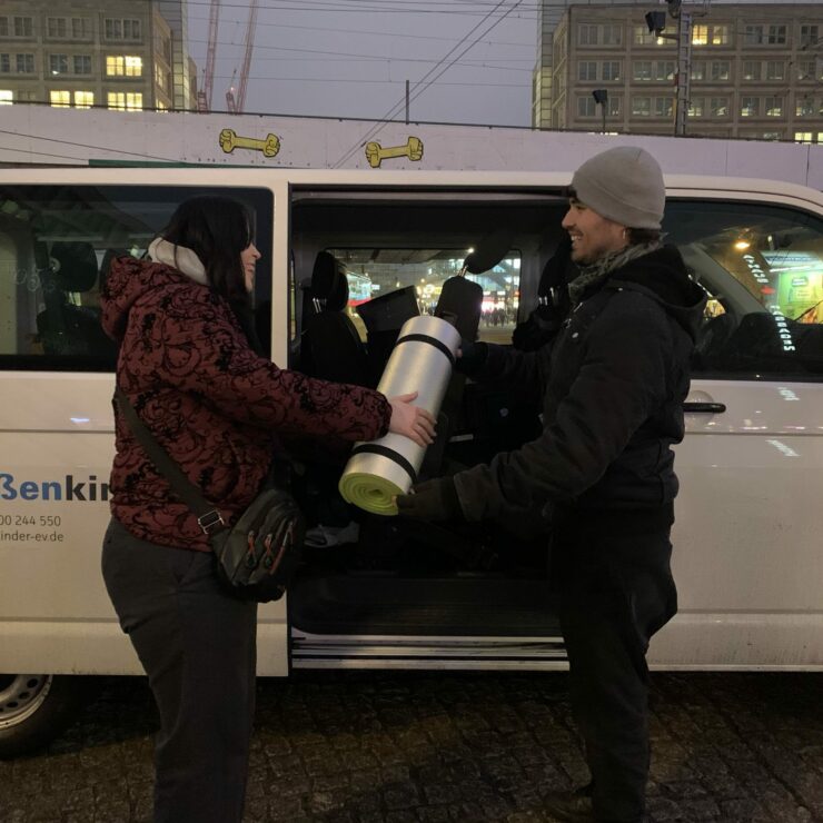 Zwei Personen stehen bei Dunkelheit vor einem weißen Kleinbus mit der Aufschrift „Straßenkinder e. V.“. Eine Person überreicht der anderen eine zusammengerollte Isomatte mit Schlafsack. Im Hintergrund sind beleuchtete Gebäude und Straßenlichter einer Stadt zu sehen.
