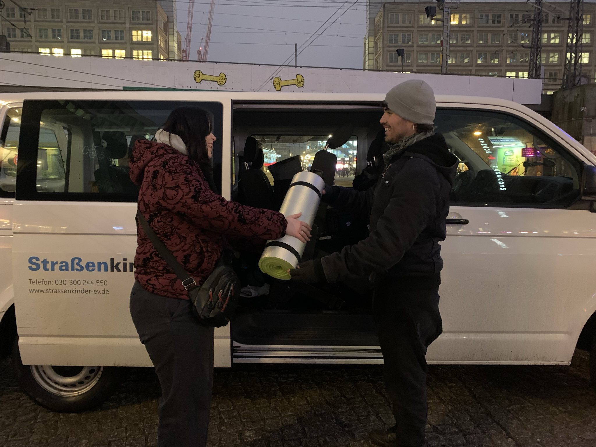 Zwei Personen stehen bei Dunkelheit vor einem weißen Kleinbus mit der Aufschrift „Straßenkinder e. V.“. Eine Person überreicht der anderen eine zusammengerollte Isomatte mit Schlafsack. Im Hintergrund sind beleuchtete Gebäude und Straßenlichter einer Stadt zu sehen.