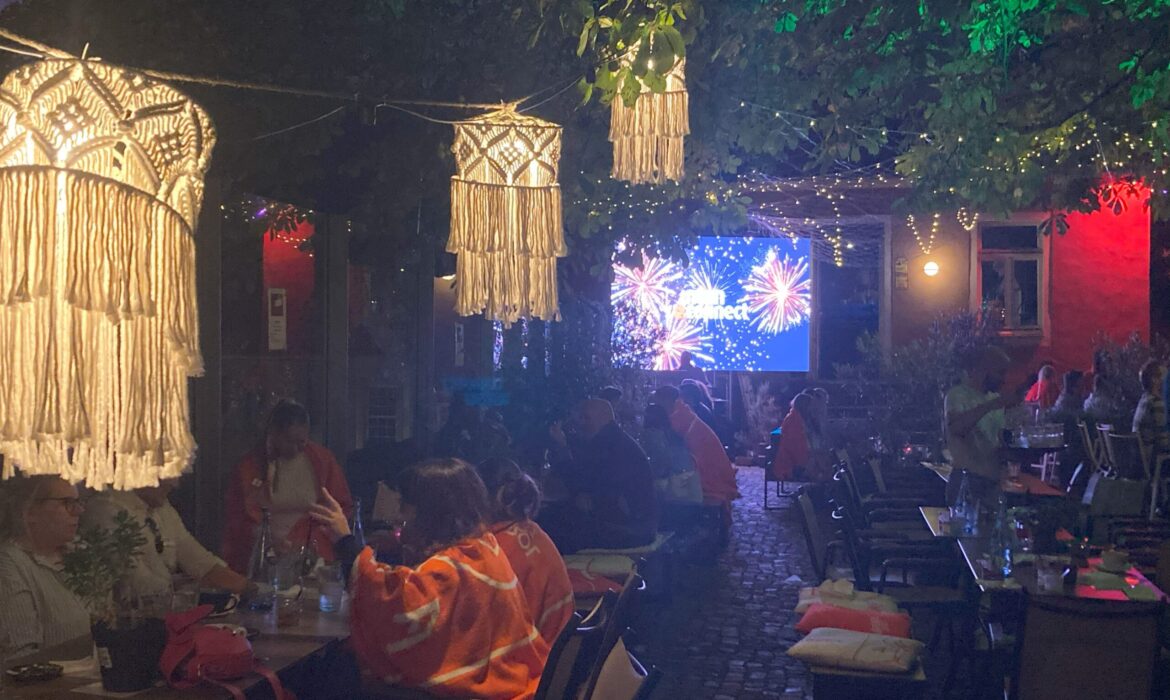 Mitarbeiter der intan group bei einem Sommerabend auf dem Heger Tor. Die Atmosphäre wird bestimmt durch beleuchtete Bäume und Leuchtmittel. Im HIntergrund ist eine Leinwand, die ein Feuerwerk zeigt.