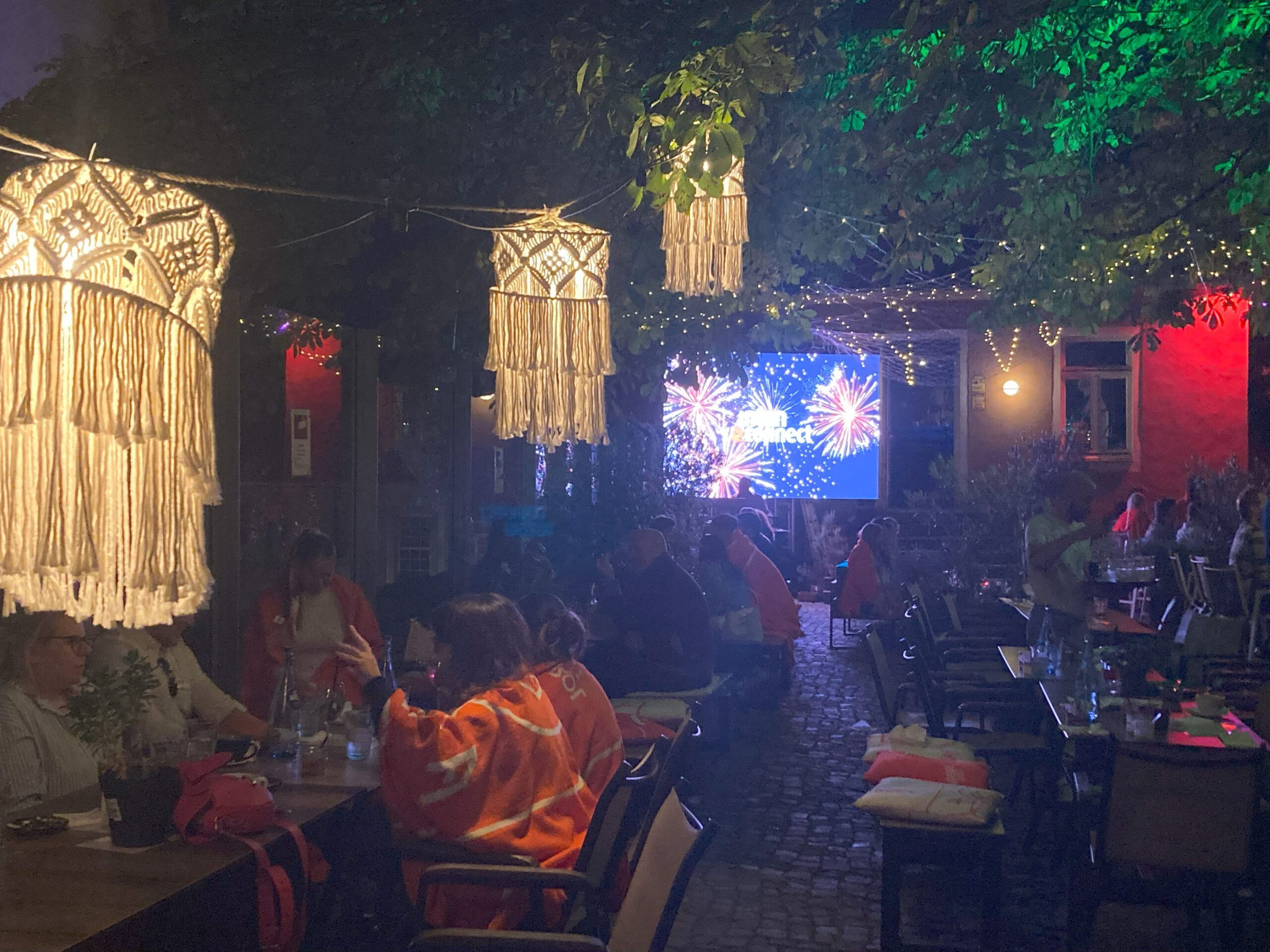 Mitarbeiter der intan group bei einem Sommerabend auf dem Heger Tor. Die Atmosphäre wird bestimmt durch beleuchtete Bäume und Leuchtmittel. Im HIntergrund ist eine Leinwand, die ein Feuerwerk zeigt.