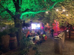 Mitarbeiter der intan group bei einem Sommerabend auf dem Heger Tor sind in Gespräüche vertieft. Die Atmosphäre wird bestimmt durch beleuchtete Bäume und Leuchtmittel. Im Hintergrund ist eine Leinwand, die ein Feuerwerk zeigt.